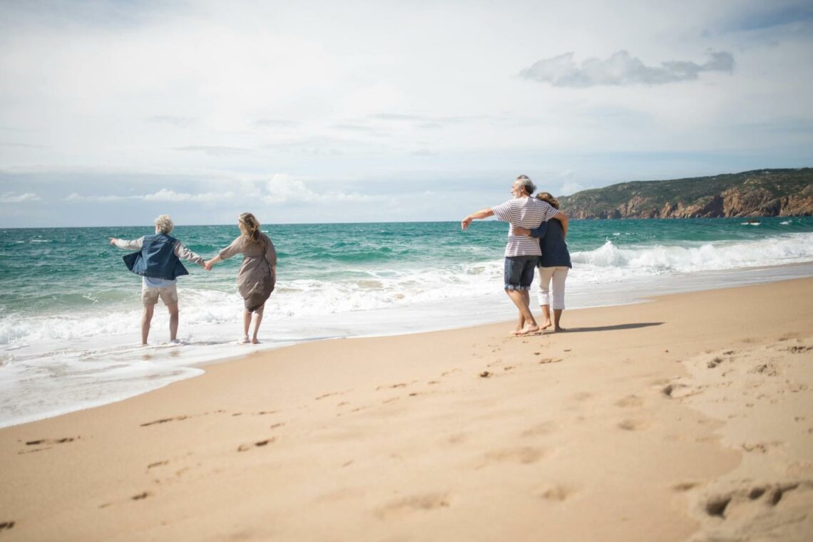 Two older couples walking along a sandy beach, holding hands and supporting each other as waves reach the shoreline on a bright, calm day.