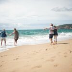Two older couples walking along a sandy beach, holding hands and supporting each other as waves reach the shoreline on a bright, calm day.