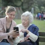Young woman helping an elderly woman use a smartphone while sitting on a park bench