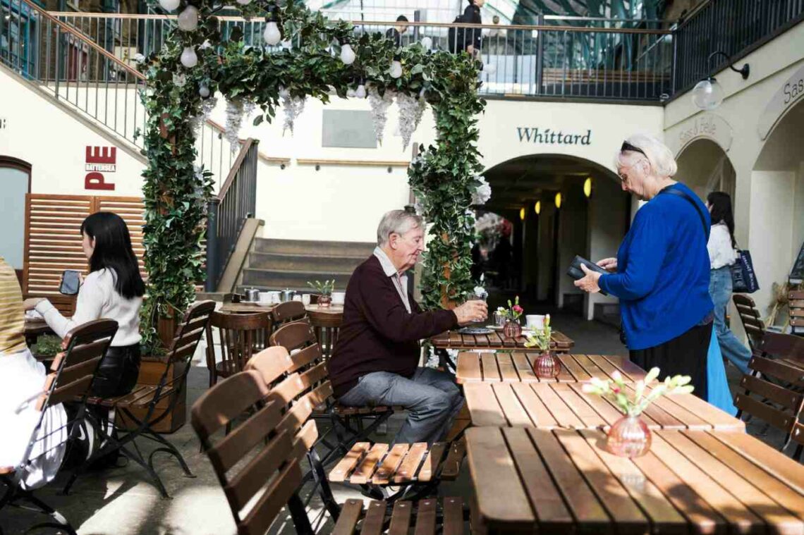 Older man sitting at an outdoor café table in London while an older woman stands beside him holding a wallet, both engaged in conversation under a leafy archway, with other people seated nearby