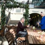 Older man sitting at an outdoor café table in London while an older woman stands beside him holding a wallet, both engaged in conversation under a leafy archway, with other people seated nearby