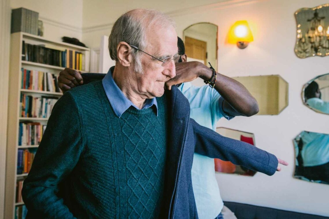 An elderly man wearing glasses and a blue sweater stands indoors while another person helps him put on a jacket. A bookshelf and wall mirrors are visible in the softly lit background.