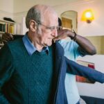 An elderly man wearing glasses and a blue sweater stands indoors while another person helps him put on a jacket. A bookshelf and wall mirrors are visible in the softly lit background.