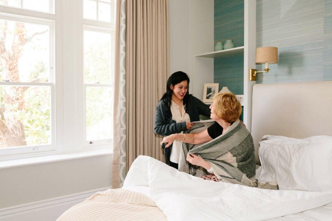 A caregiver helps an elderly woman sit up in bed, gently wrapping a blanket around her shoulders in a bright, comfortable bedroom with large windows.