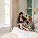 A caregiver helps an elderly woman sit up in bed, gently wrapping a blanket around her shoulders in a bright, comfortable bedroom with large windows.