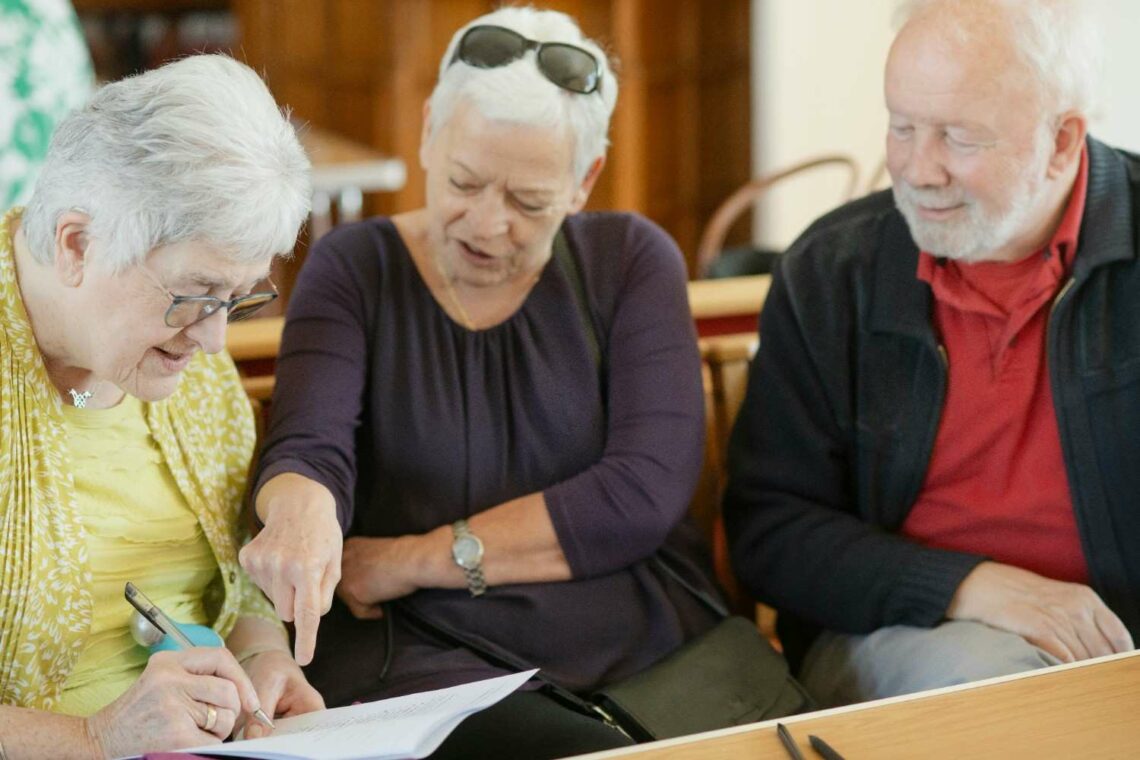 Three older adults seated together, one woman writing on a document while another points at the page
