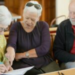 Three older adults seated together, one woman writing on a document while another points at the page