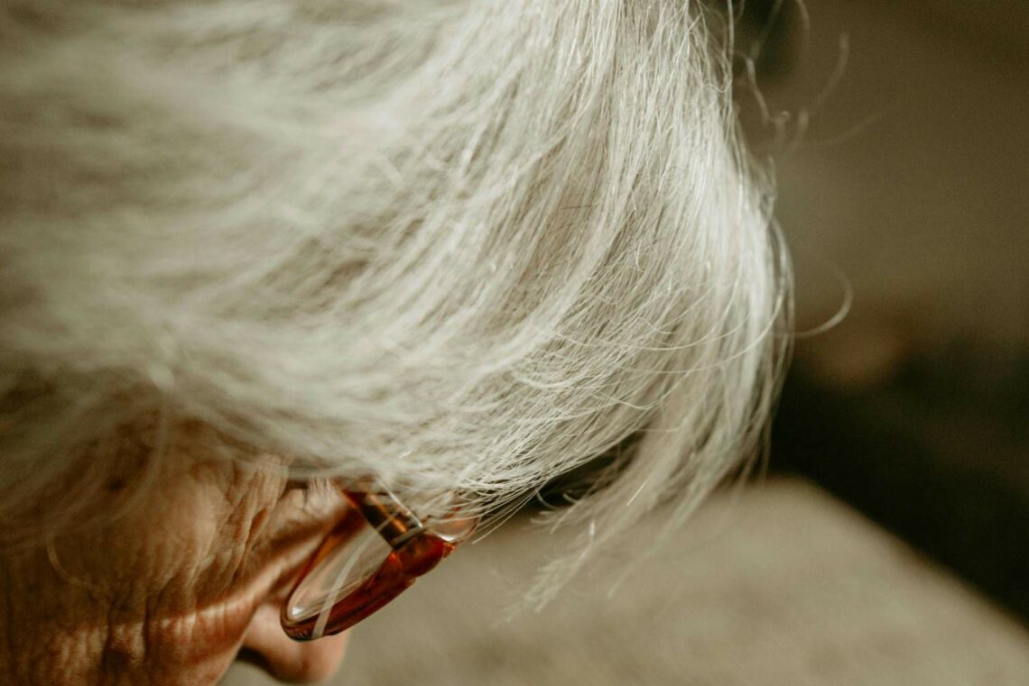 A senior woman with glasses and white hair, looking thoughtfully while sitting in a well-lit room.