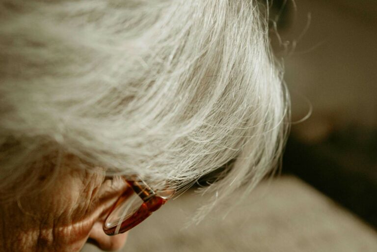 A senior woman with glasses and white hair, looking thoughtfully while sitting in a well-lit room.