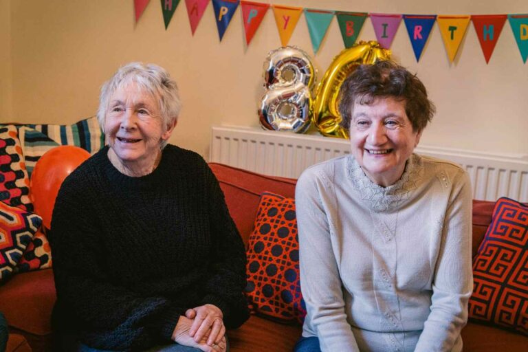 Two older women sit on a couch surrounded by colorful balloons and birthday decorations, celebrating a special occasion.