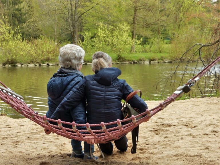 Older woman sitting beside a caregiver on a hammock by a lake, both wearing jackets and sharing a quiet moment outdoors.
