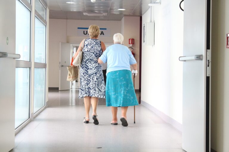 An elderly woman using a walking stick is supported by a companion as they walk down a hospital corridor after discharge.