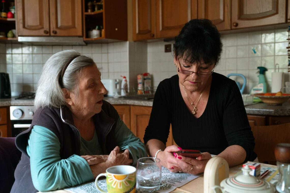 An older woman and a younger woman sit together at a kitchen table, engaged in conversation and sharing a meal.