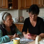 An older woman and a younger woman sit together at a kitchen table, engaged in conversation and sharing a meal.