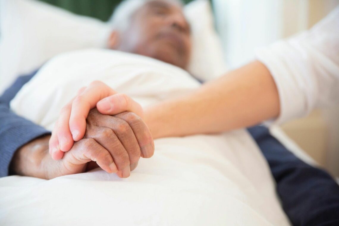 A younger person holding the hand of an older man lying in a bed at home