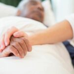 A younger person holding the hand of an older man lying in a bed at home