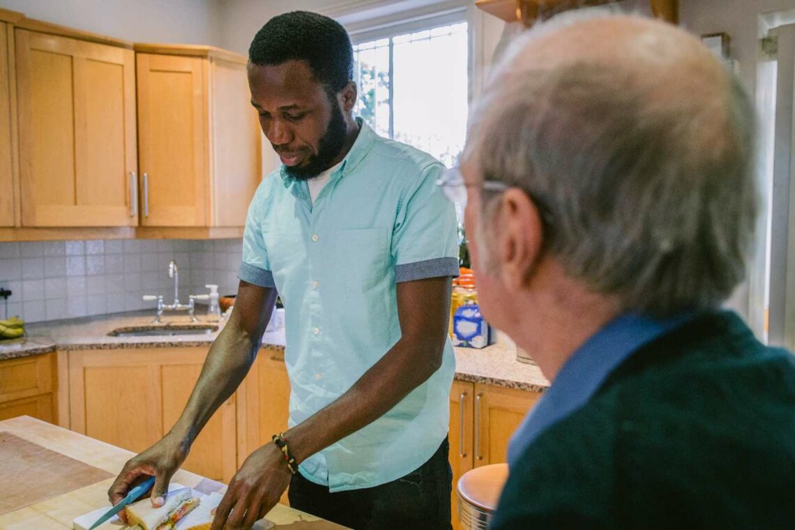 A carer preparing food in a home kitchen while an older man looks on
