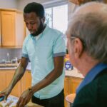 A carer preparing food in a home kitchen while an older man looks on