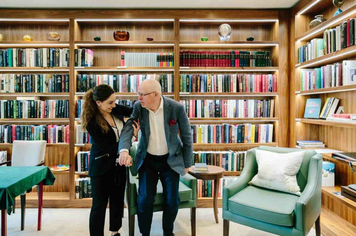 A woman supports an elderly man with a cane as they walk together in a warmly lit library lined with wooden bookshelves filled with colorful books. Two green armchairs and a small table sit nearby, creating a cozy reading space.