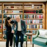 A woman supports an elderly man with a cane as they walk together in a warmly lit library lined with wooden bookshelves filled with colorful books. Two green armchairs and a small table sit nearby, creating a cozy reading space.