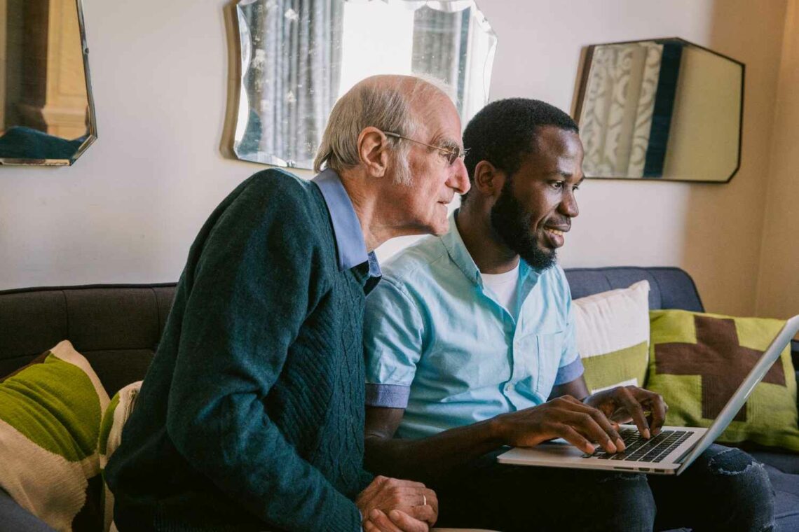 Elderly man and younger man sitting together on a sofa, smiling and looking at a laptop screen, suggesting companionship and shared learning or support.
