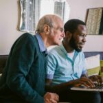 Elderly man and younger man sitting together on a sofa, smiling and looking at a laptop screen, suggesting companionship and shared learning or support.