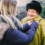 Smiling elderly woman wearing a green coat and black hat being helped by a younger woman outdoors, conveying warmth, care, and companionship.