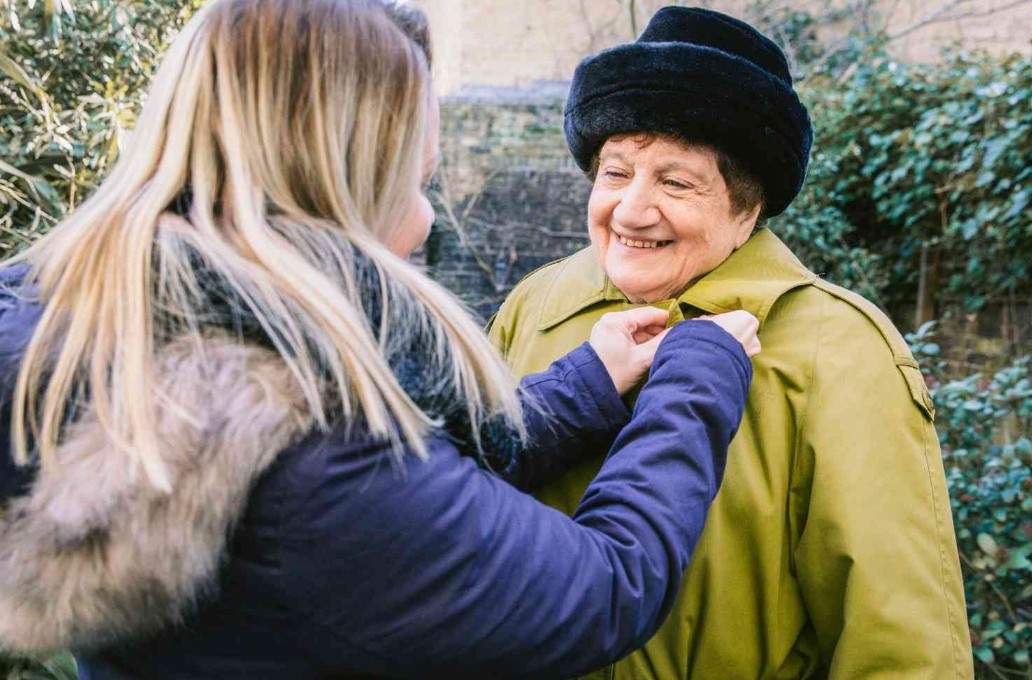 Smiling elderly woman wearing a green coat and black hat being helped by a younger woman outdoors, conveying warmth, care, and companionship.