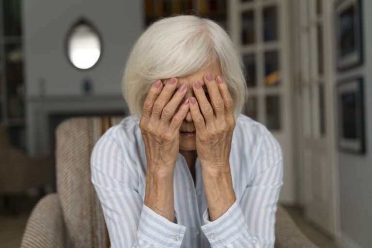 Elderly woman sitting with her head in her hands, expressing sadness or distress in a quiet home setting.