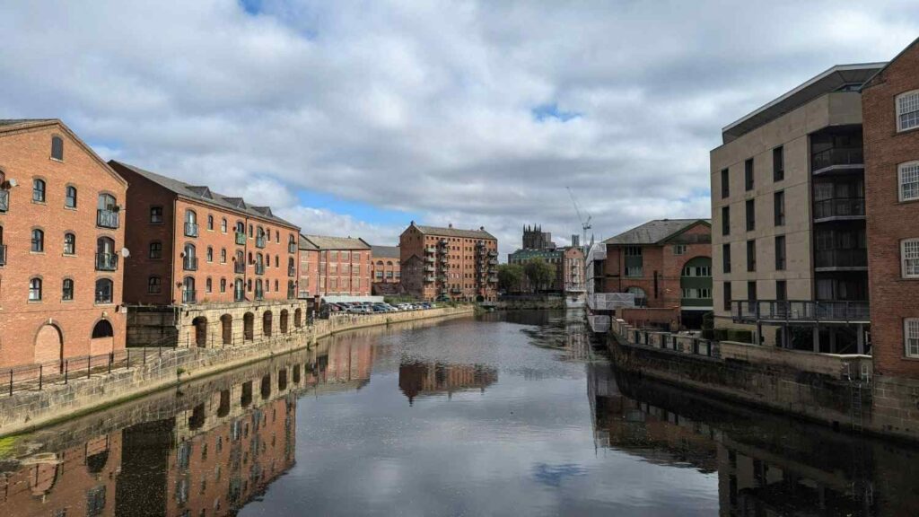 View of Leeds city centre along the River Aire, with converted red-brick warehouses and modern apartments lining the waterfront under a cloudy sky, reflected in the still water.