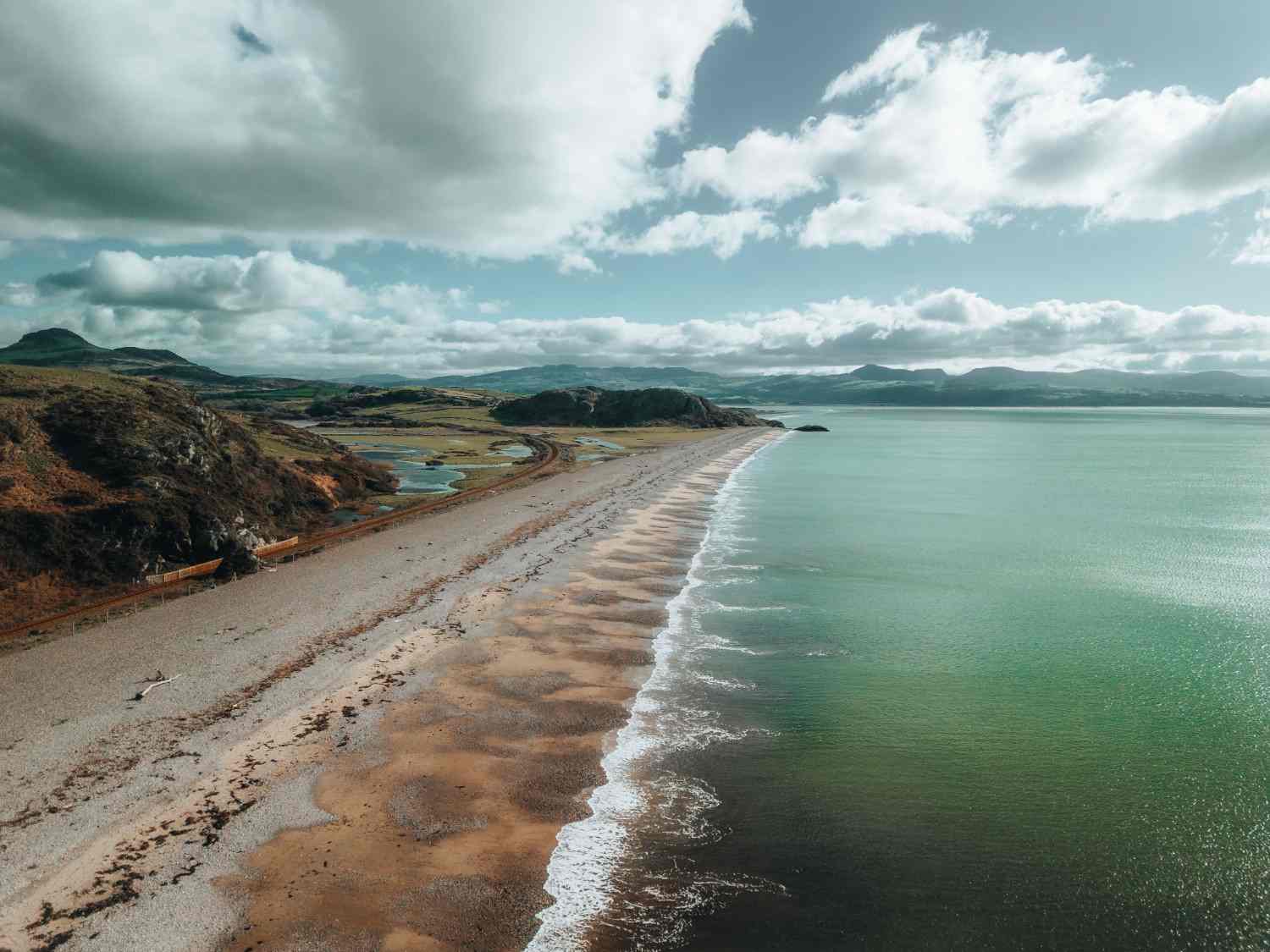 Aerial view showcasing a sandy beach bordered by lush green mountains under a clear blue sky.