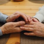 Close-up of two elderly people holding hands on a table, symbolising comfort, care, and emotional support in dementia care.