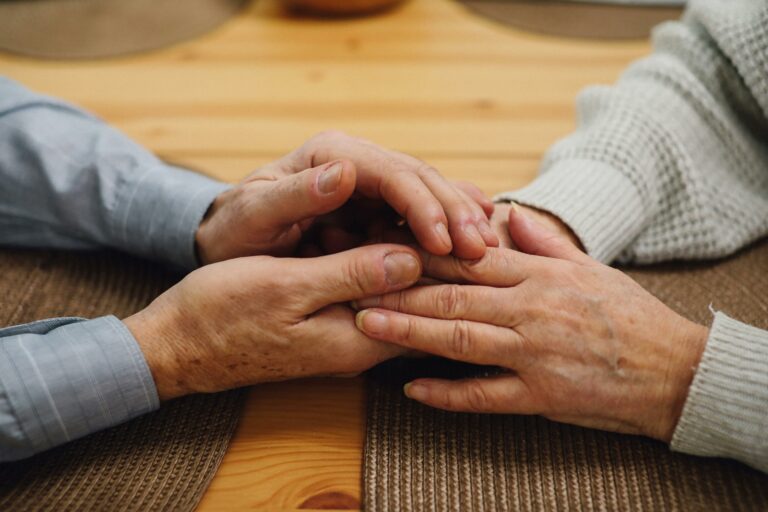 Close-up of two elderly people holding hands on a table, symbolising comfort, care, and emotional support in dementia care.
