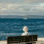 An elderly person with short white hair sits alone on a wooden bench facing the sea, watching the waves and a distant boat under a clear sky.