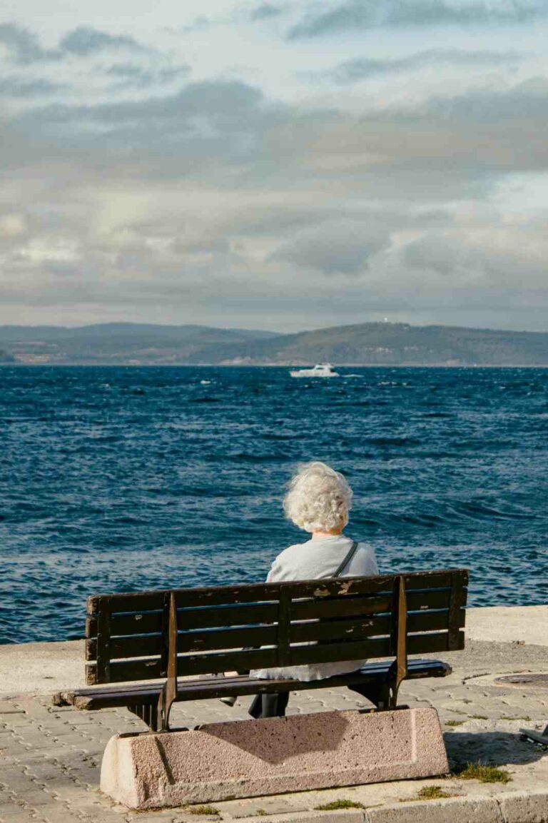 An elderly person with short white hair sits alone on a wooden bench facing the sea, watching the waves and a distant boat under a clear sky.