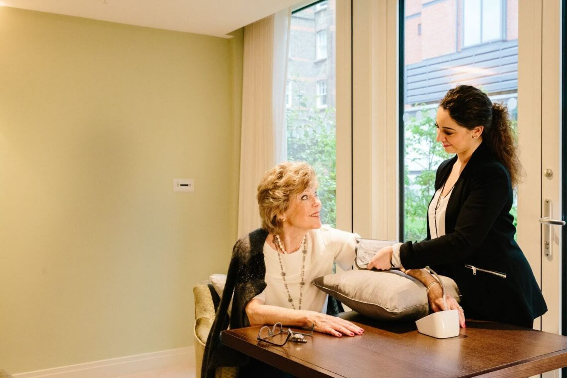 A smiling caregiver checks the blood pressure of an older woman seated at a table. The woman rests her arm on a cushion, and they share a warm, friendly interaction in a bright room with large windows.