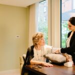 A smiling caregiver checks the blood pressure of an older woman seated at a table. The woman rests her arm on a cushion, and they share a warm, friendly interaction in a bright room with large windows.