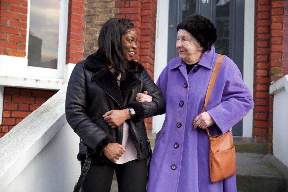 Care worker supporting an elderly woman as they walk together outside a home.