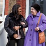 Care worker supporting an elderly woman as they walk together outside a home.