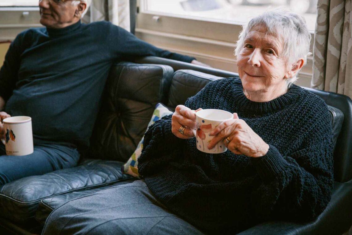 Elderly woman smiling while holding a mug, sitting on a sofa next to an elderly man, in a bright living room with natural light from a window.