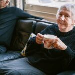 Elderly woman smiling while holding a mug, sitting on a sofa next to an elderly man, in a bright living room with natural light from a window.