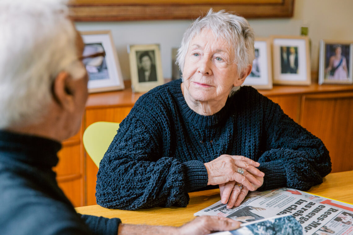 Elderly woman sitting at a table and listening attentively to another person, conveying empathy, connection, and meaningful conversation in a home setting.