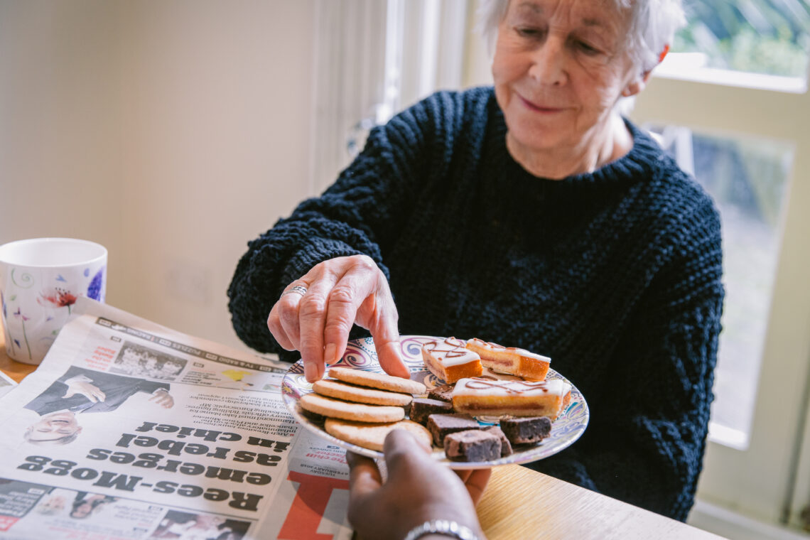 Carer offering biscuits to an elderly woman, showing comfort and companionship during mealtime.