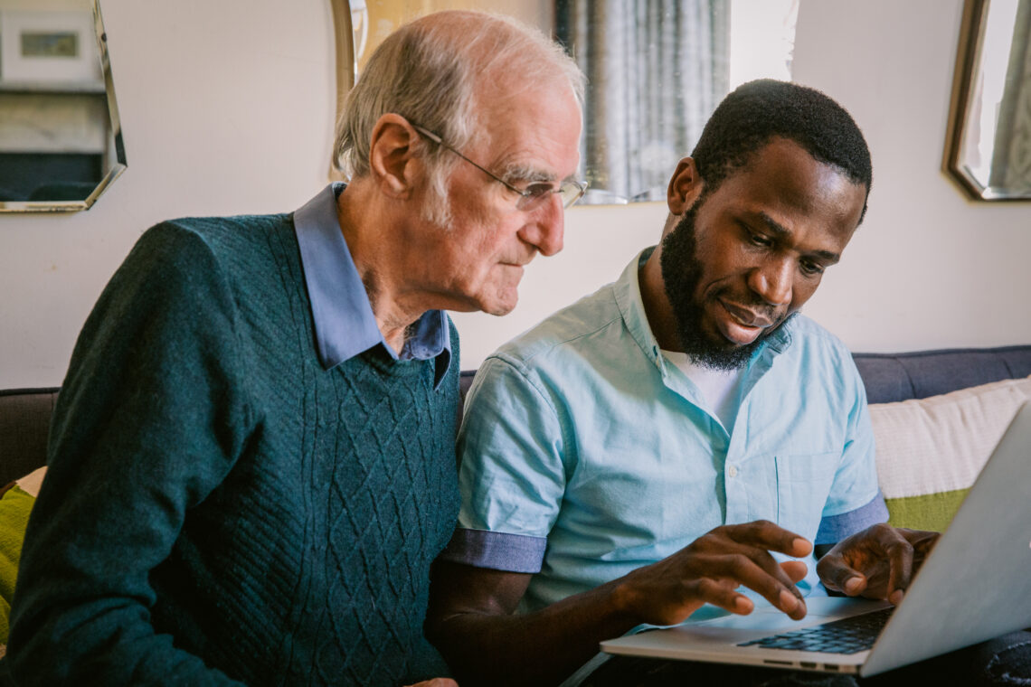Younger man assisting an older man on a laptop, showing patient support and digital inclusion in elderly care.