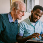 Younger man assisting an older man on a laptop, showing patient support and digital inclusion in elderly care.