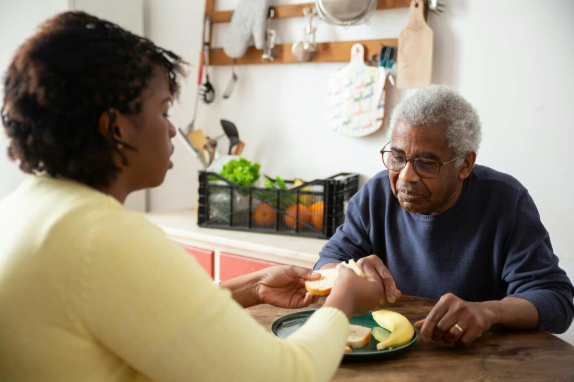 Elderly man sitting at a kitchen table being helped with a meal by a caregiver, showing support and companionship in a home setting.