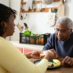 Elderly man sitting at a kitchen table being helped with a meal by a caregiver, showing support and companionship in a home setting.
