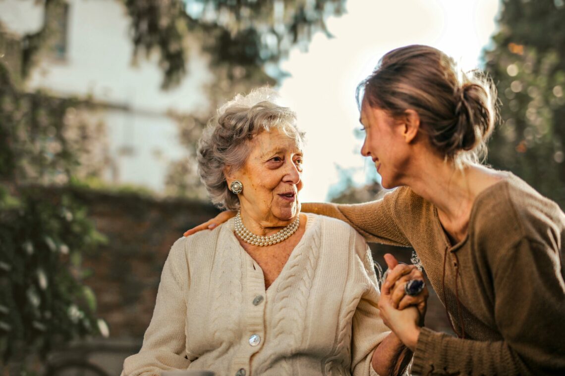 Smiling elderly woman sitting outdoors and holding hands with a younger caregiver, highlighting warmth and companionship in elderly care.