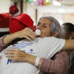 An older woman embraces someone wearing a Santa hat during a festive gathering, highlighting warmth, connection and holiday companionship.