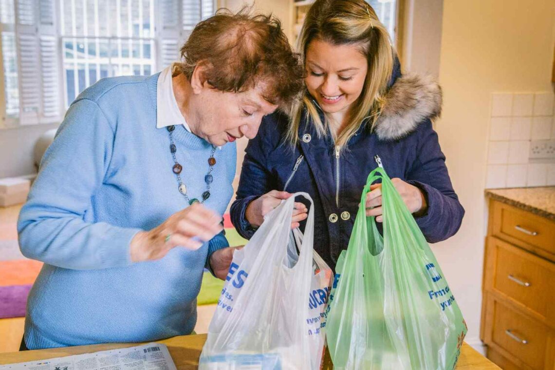 Visiting carer supporting an older woman at home while unpacking shopping in the kitchen.